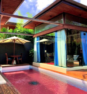 A modern outdoor pool area with a wooden deck, two umbrellas, and a glass-enclosed space with blue curtains. - Kiss Villas Bali (Seminyak)