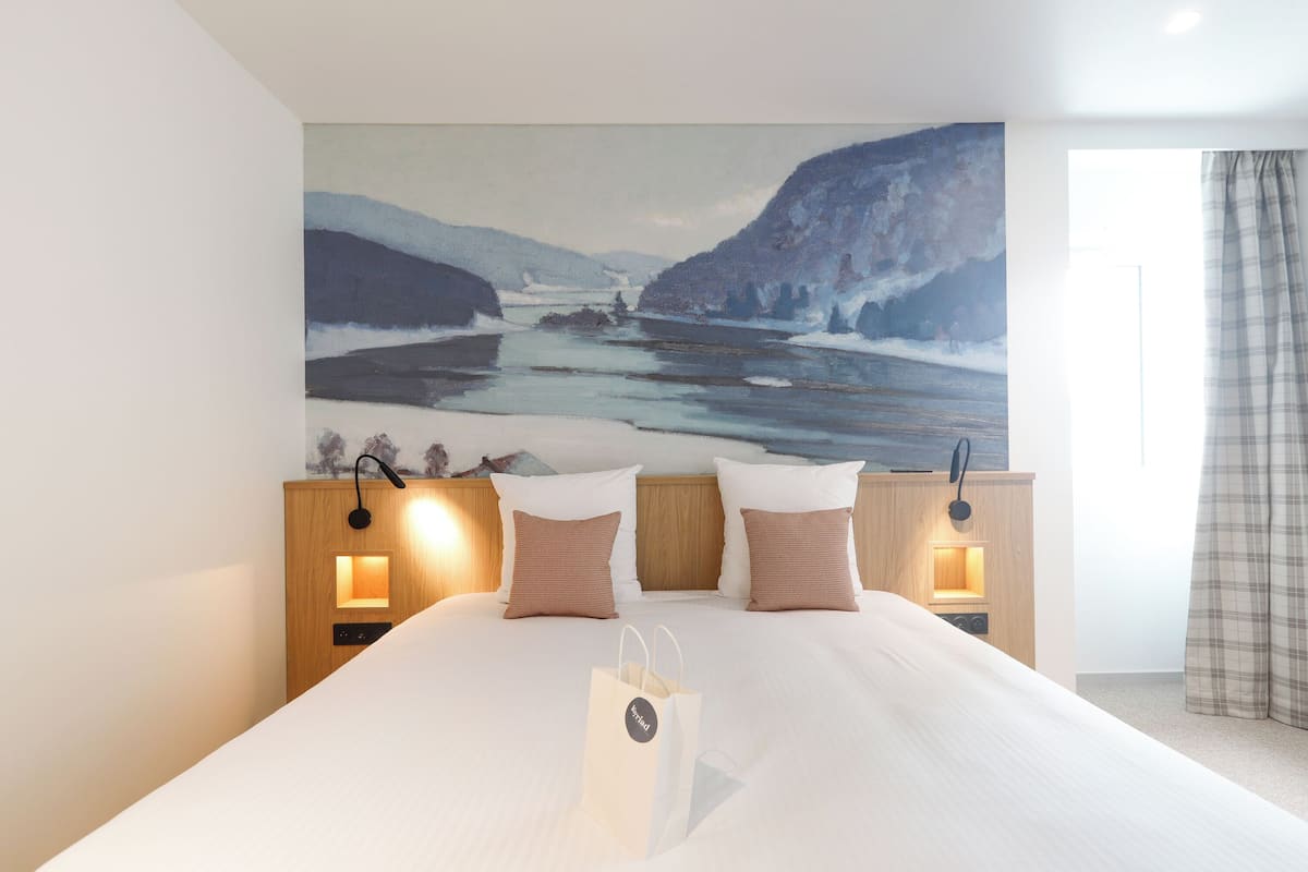 Quarto de hotel com uma cama grande, duas luminárias de cabeceira, uma mesa de cabeceira e um quadro com paisagem.