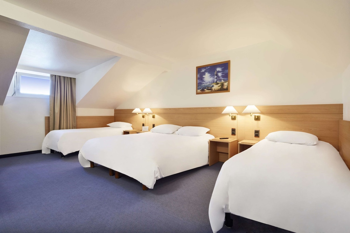 Habitación de hotel con tres camas dispuestas en fila, un cuadro de un faro en la pared y un tragaluz arriba.