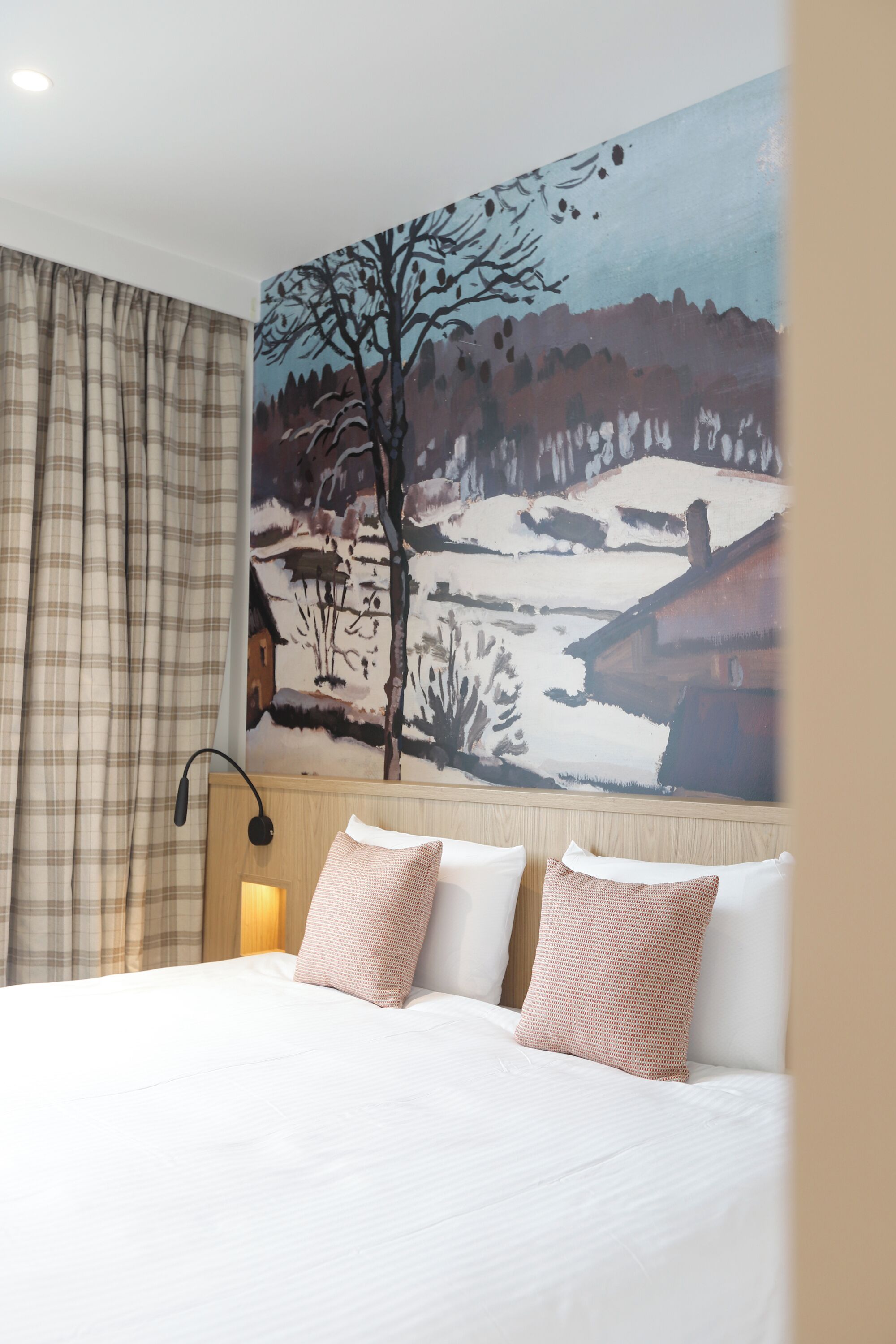 A hotel room with a bed, a headboard with a painting, a lamp, and a window with curtains.
