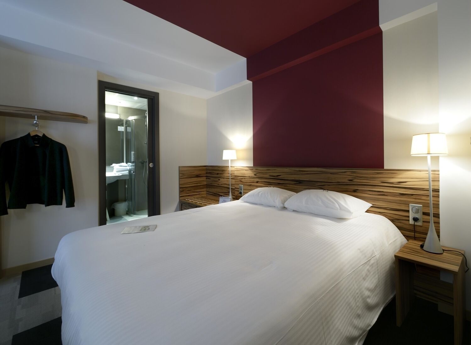 Et hotelværelse med en seng, sengeborde med lamper, en træsenggavl, et knagered og et spejl.