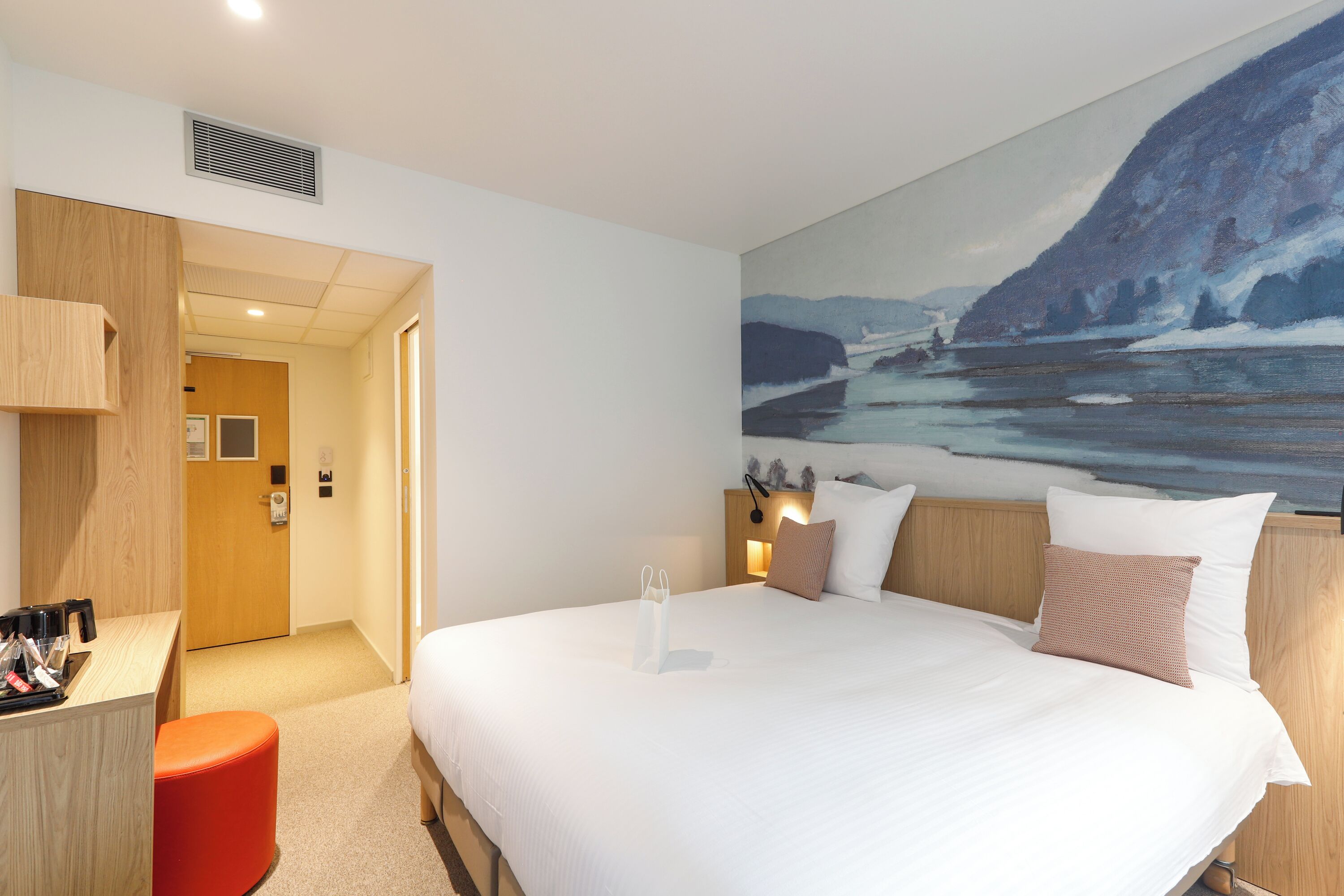 A hotel room with a bed, a desk, a chair, a painting on the wall, and a door.