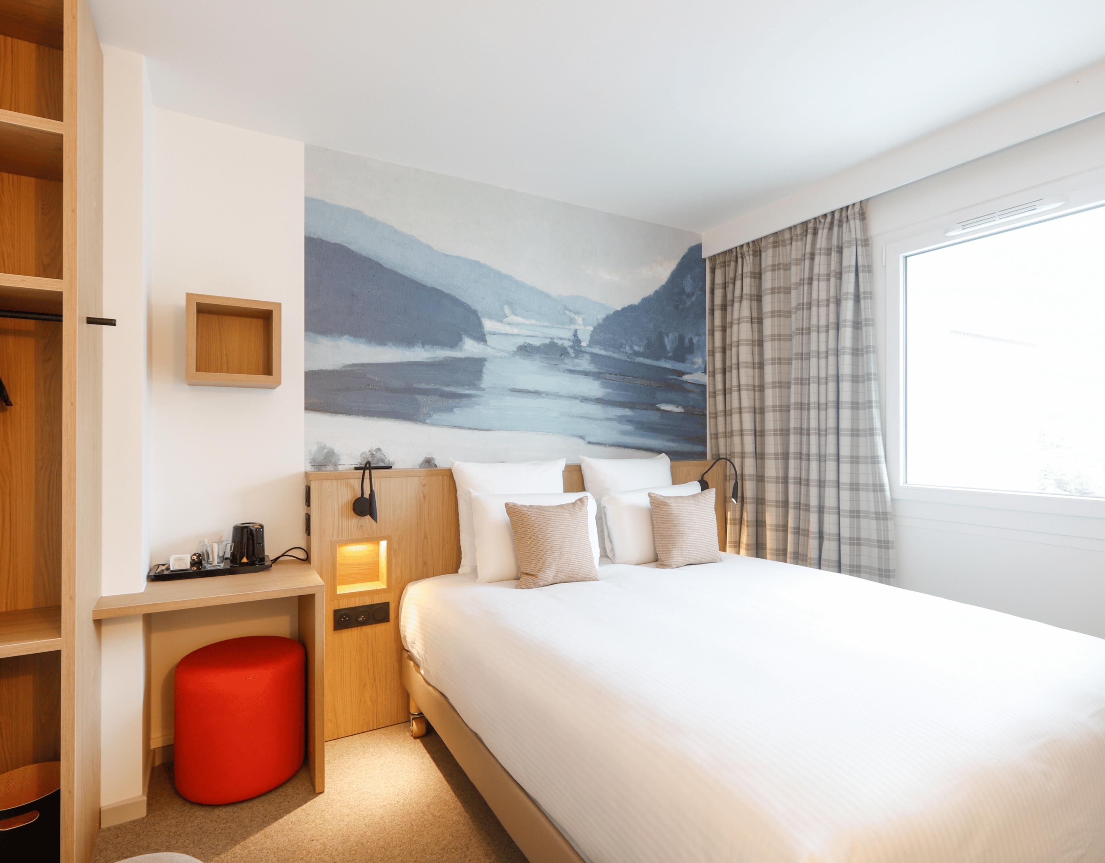 Habitación de hotel con cama, un reposapiés rojo, una mesita de noche con lámpara, estante montado en la pared y un ventanal con cortinas.