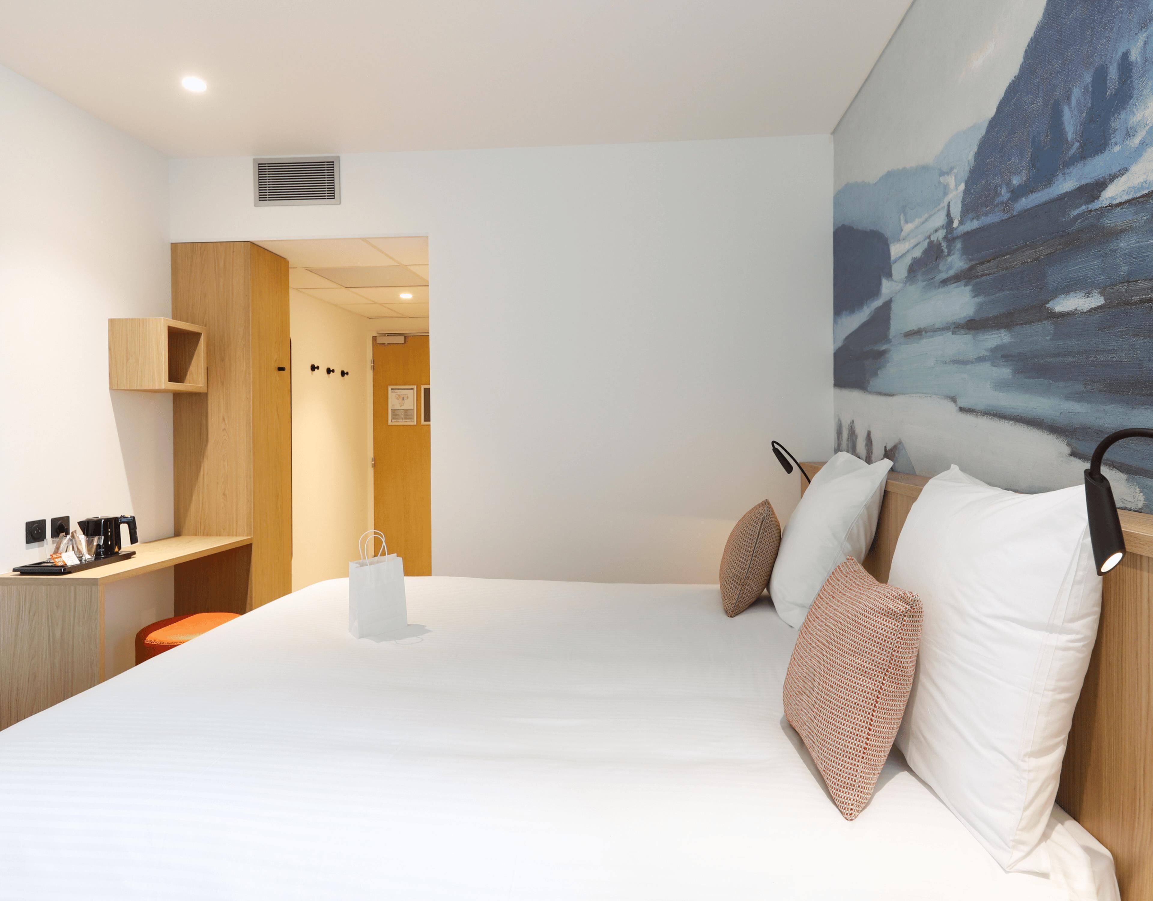 A modern hotel room with a large bed, a desk with a chair, a painting on the wall, and a door leading to another room.