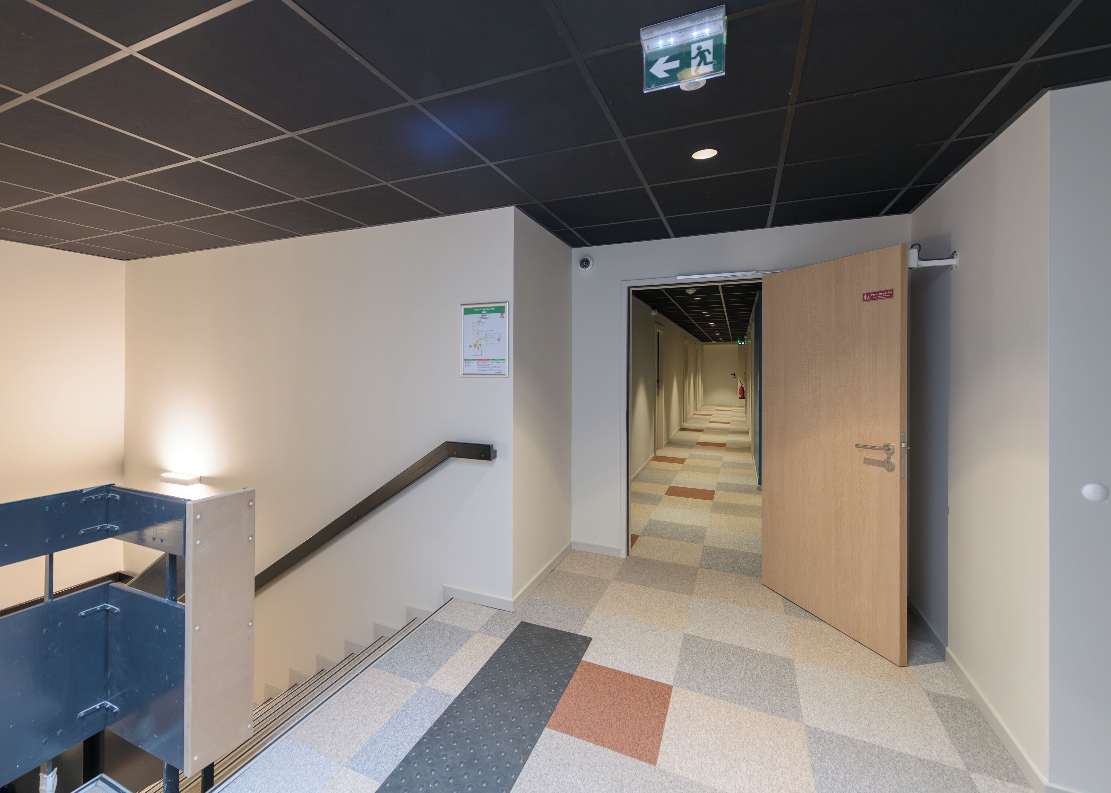 Un couloir avec un sol à carreaux, un panneau indiquant la sortie de secours et une porte dotée d’un trou de serrure.