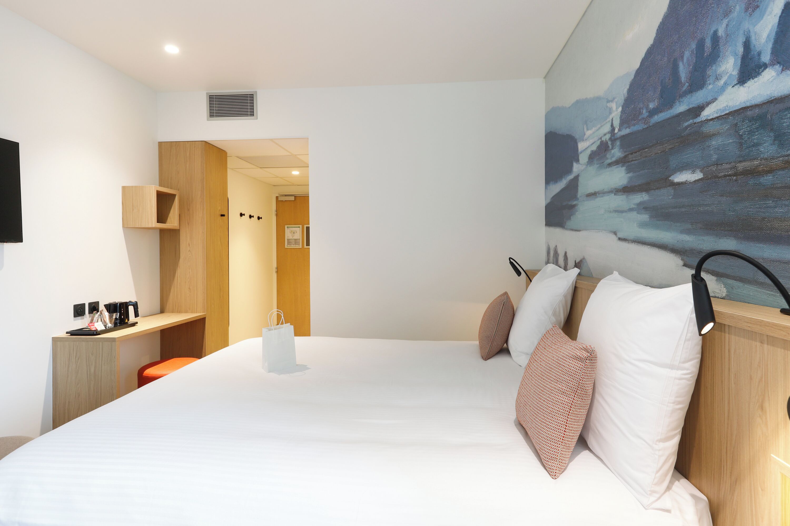 A modern hotel room with a large bed, a desk with a chair, a painting on the wall, and a view of the outdoors.