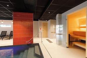 A modern indoor pool area with a red-tiled wall, a wooden sauna, and seating areas.