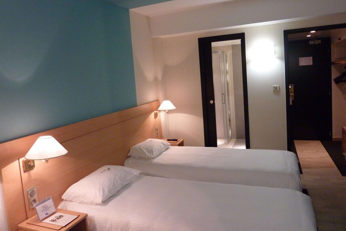 Una habitación de hotel con dos camas, una mesita de noche, una lámpara y una puerta que da a otra habitación.