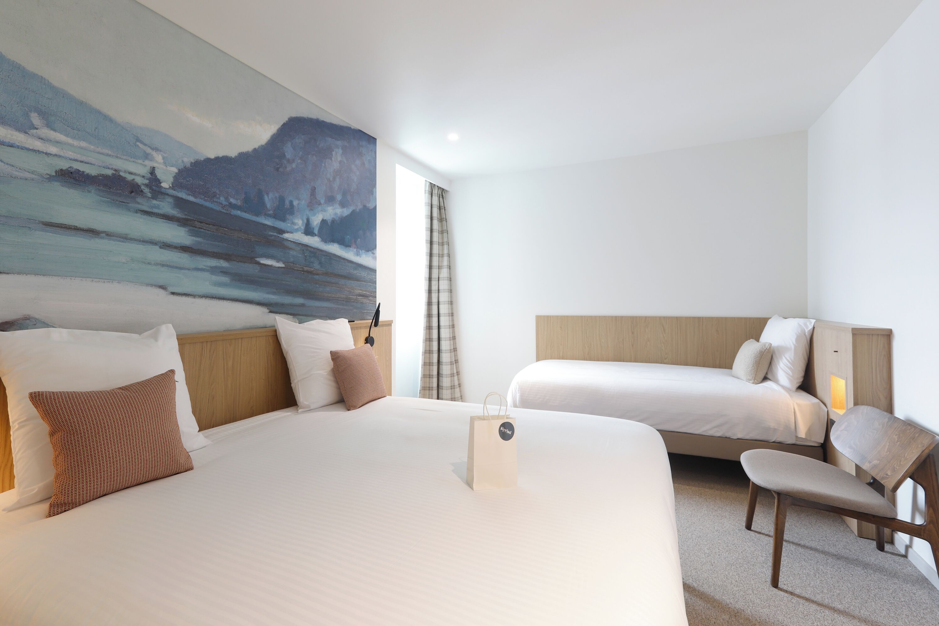 Une chambre d’hôtel avec deux lits, un tableau représentant un paysage de montagne, une fenêtre avec des rideaux et une table de chevet avec une lampe.
