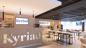Moderni hotellin aula, jossa on vastaanottotiski, jonka kyltissä lukee ”Kyriad”, ruokailutila pöytineen ja tuoleineen sekä suuri näyttö, jossa näkyy hotellin nimi.