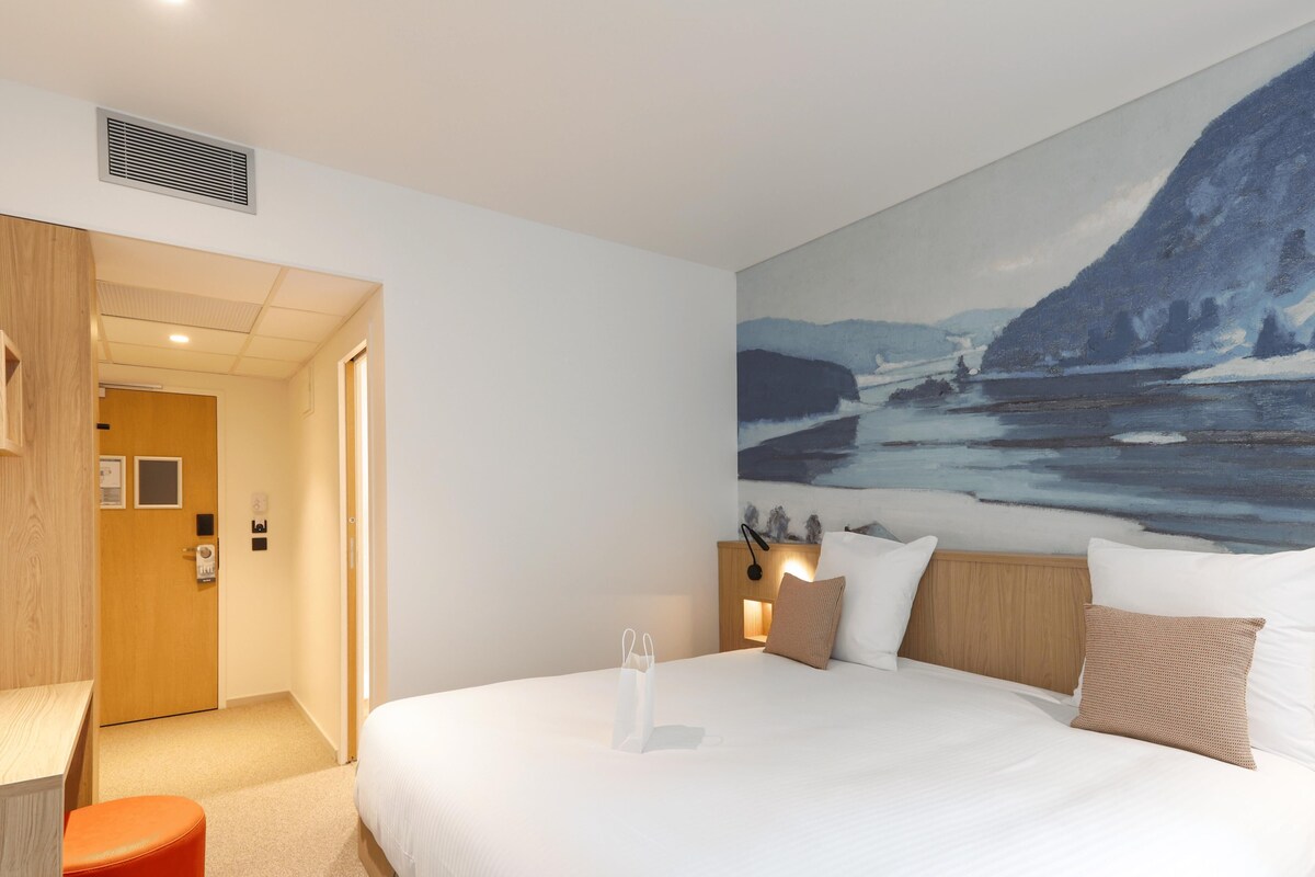Habitación de hotel con cama, un cuadro de un paisaje nevado, cabecero de madera y un reposapiés rojo.