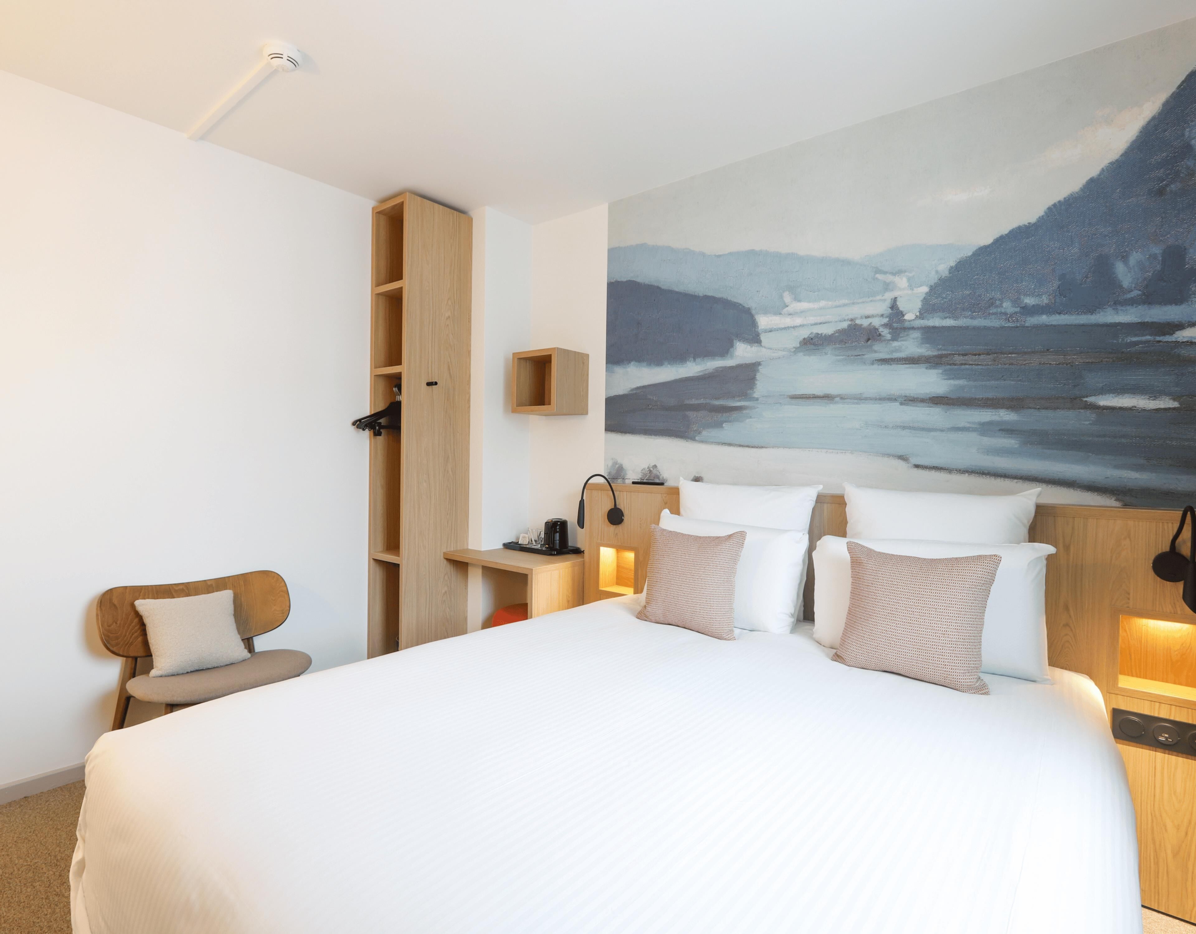 Quarto de hotel moderno com uma cama grande, guarda-roupa de madeira, escrivaninha com luminária e uma pintura de uma paisagem costeira.