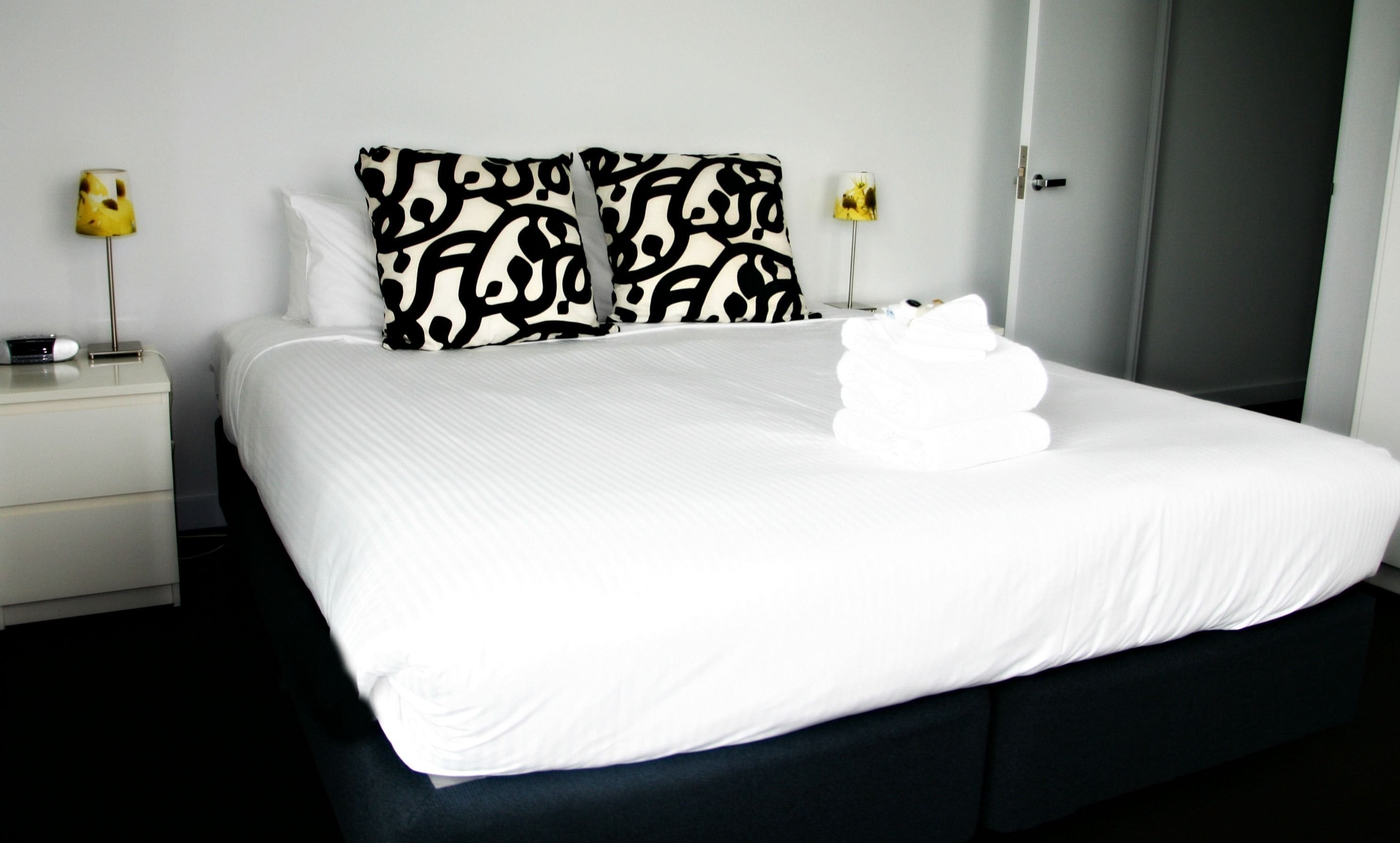 Un lit bien fait, avec du linge de lit blanc et un coussin à motifs noir et blanc, une table de chevet avec une lampe, et une porte visible au fond.