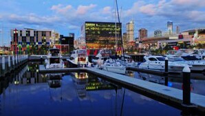Uma marina com barcos atracados, um horizonte urbano ao fundo e um céu claro.