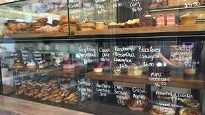 Vitrine de pastelaria com diversos produtos de panificação e sobremesas, incluindo bolos, doces e sanduíches, com preços indicados no vidro.