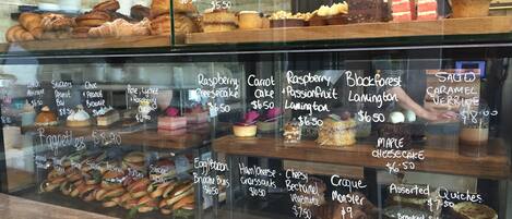 A bakery display case with various baked goods and desserts, including cakes, pastries, and sandwiches, with prices listed on the glass.