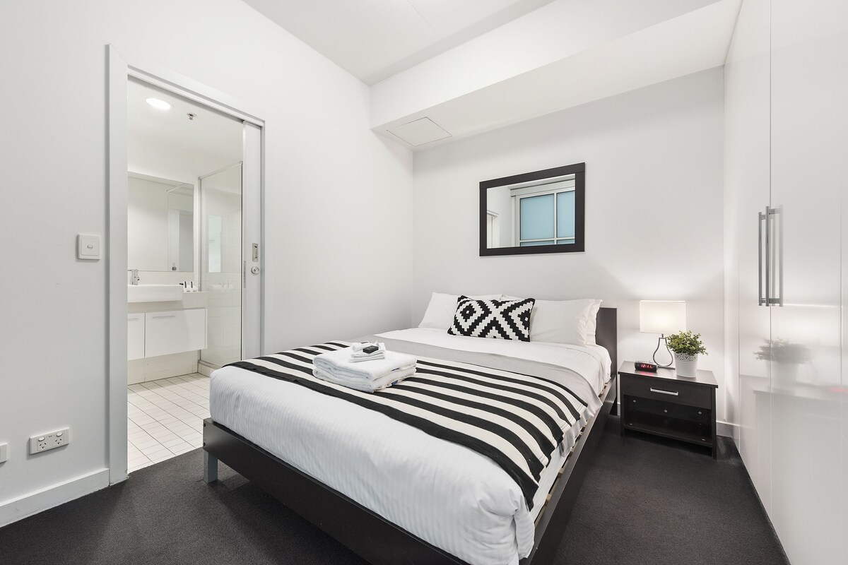 Quarto moderno com cama, mesa de cabeceira e espelho.