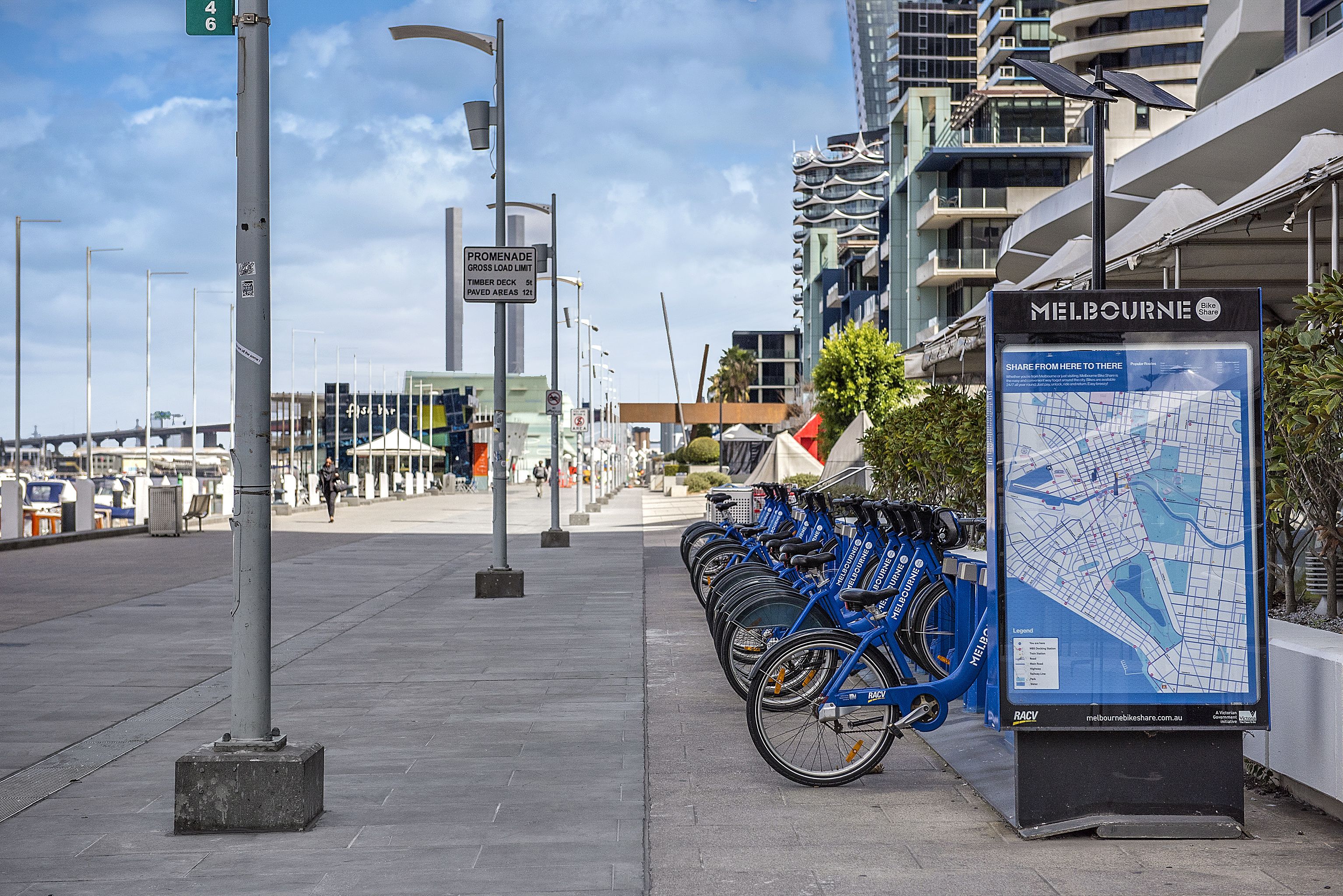 A row of blue bicycles parked on a sidewalk, a map sign, and a street sign indicating a 46 km limit.