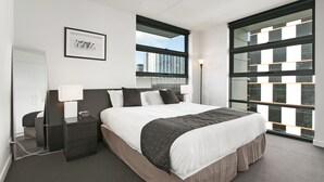 Quarto de hotel moderno com uma cama grande, mesa de cabeceira, luminária na parede e uma janela ampla com vista para a cidade.