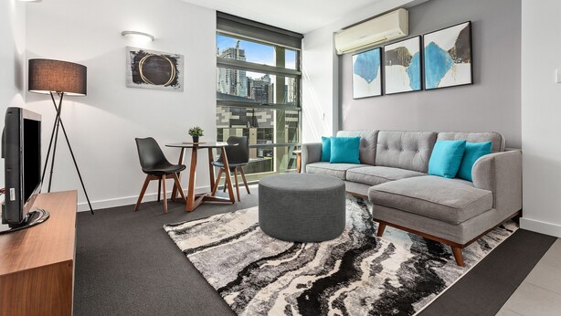 Uma sala de estar moderna com um sofá sectional cinza, um banquinho redondo, um tapete preto e branco, uma TV em um suporte de madeira e vista para a cidade através de janelas amplas.