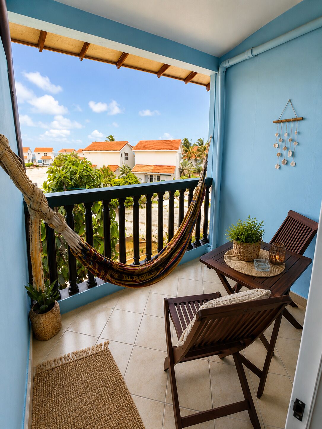 Un balcon avec un hamac, deux chaises en bois, un tapis en fibres naturelles et une vue sur les maisons et la verdure.