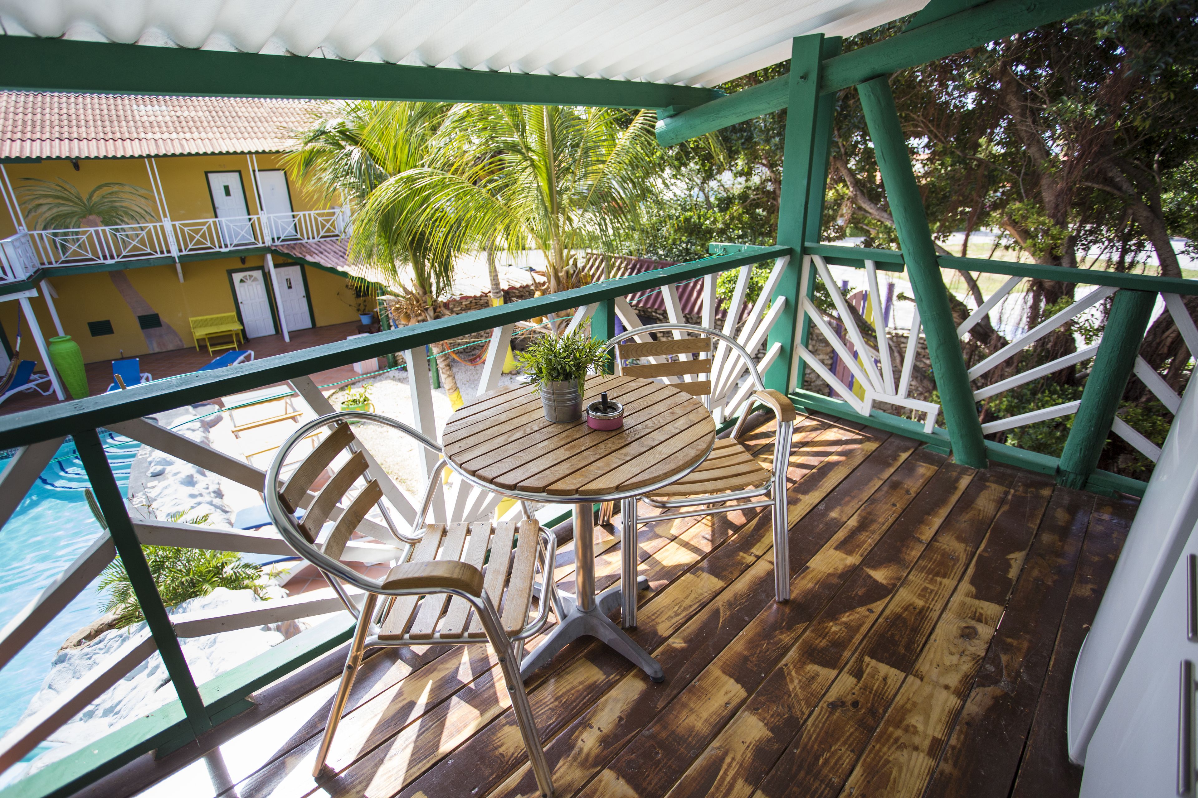 Eine Holzterrasse mit rundem Tisch und Stühlen, mit Blick auf einen Swimmingpool und ein Gebäude mit Balkon.