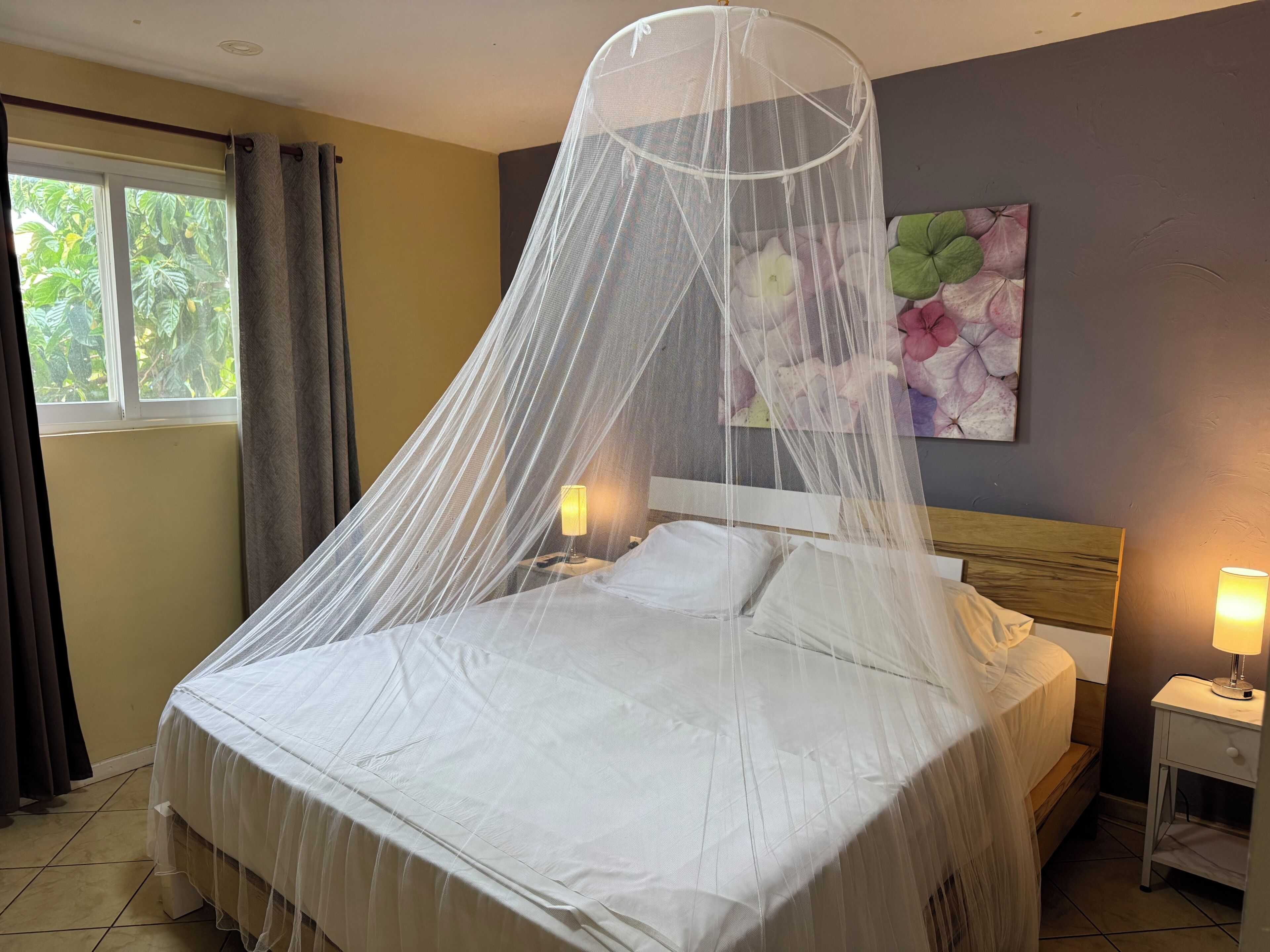 Una cama con dosel, dos lámparas de noche, una mesita de noche y un cuadro con motivos florales en la pared.