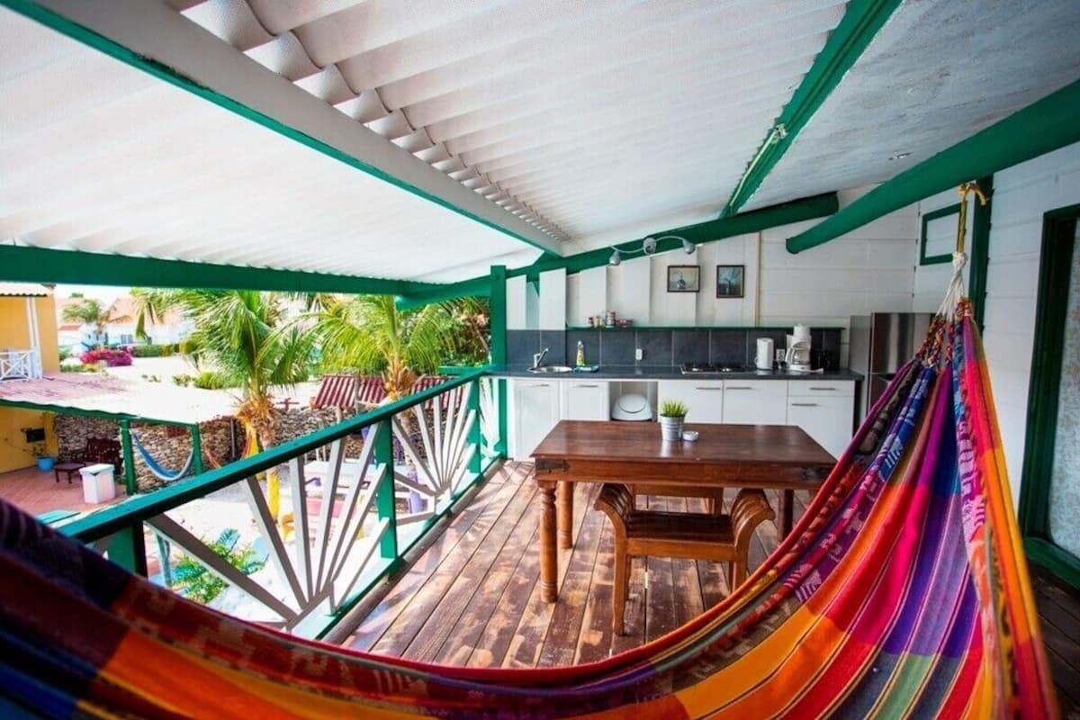 A covered outdoor area with a wooden table, a hammock, and a kitchen area with white cabinets.