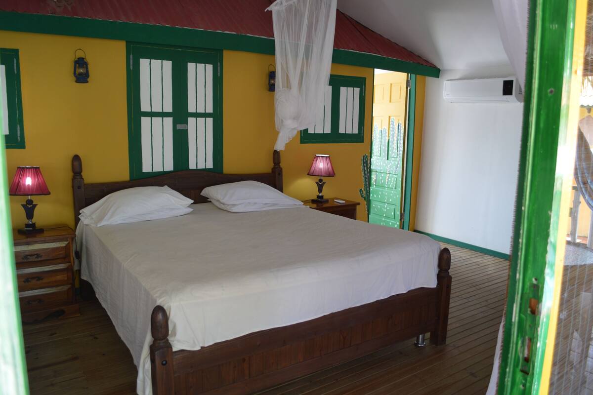 A bedroom with a wooden bed, two bedside tables, a nightstand, a lamp, a mosquito net, and a green door.