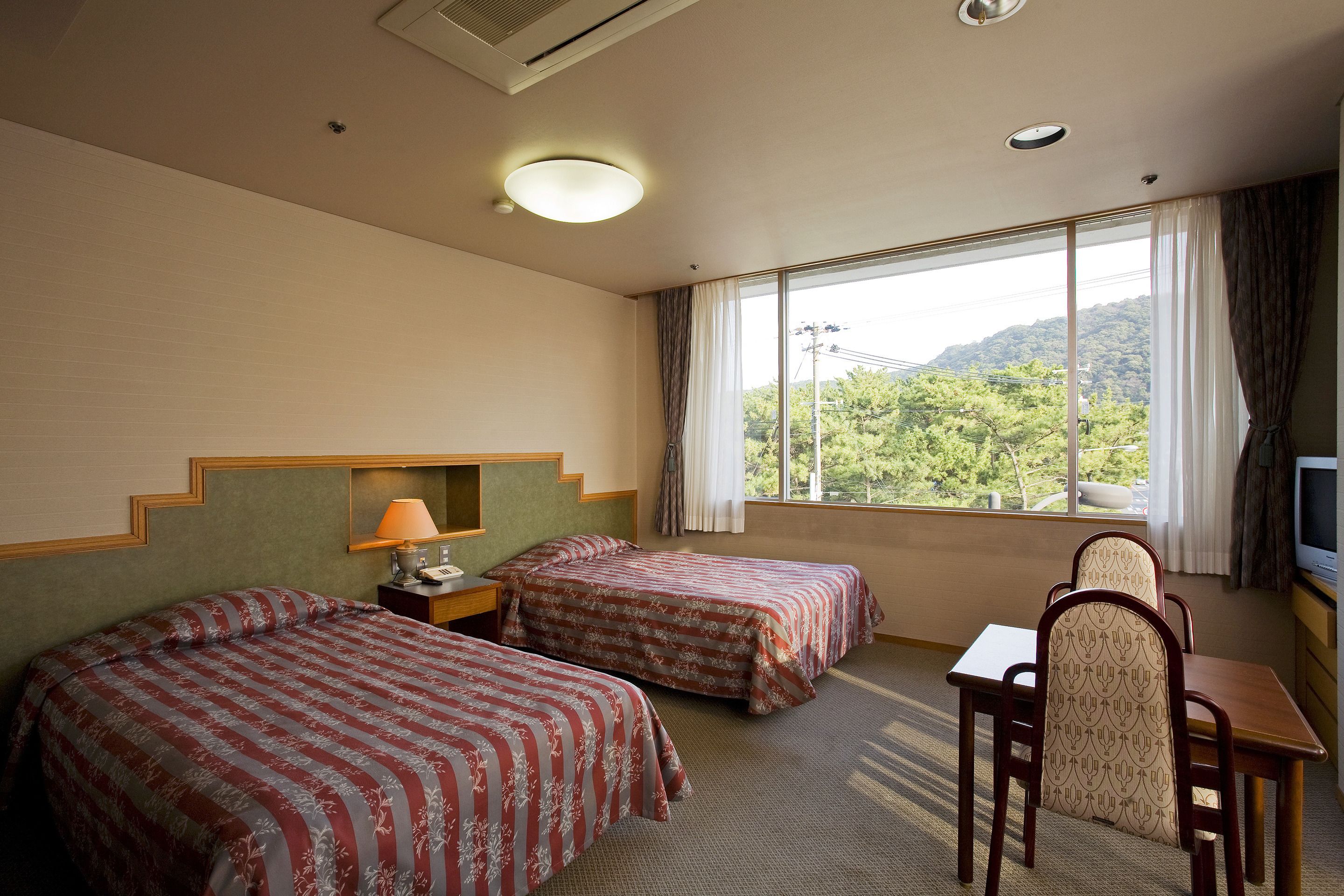 A hotel room with two beds, a desk, a chair, a television, and a window with a view of greenery.