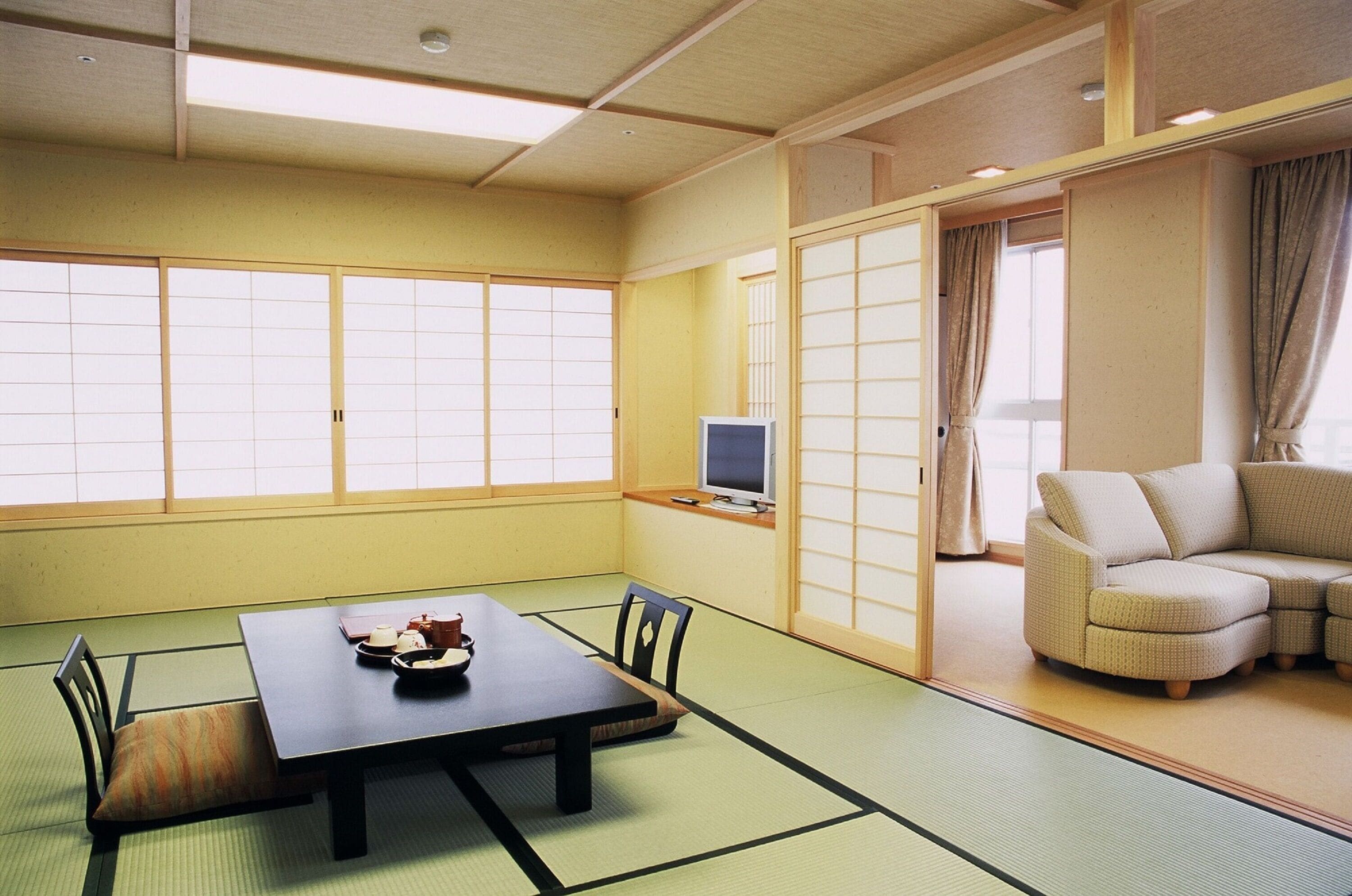 Une pièce de style japonais traditionnel, avec un sol recouvert de tatamis, une table basse en tatami, un canapé bas, des portes coulissantes en papier de riz (shoji) et un téléviseur posé sur un meuble.