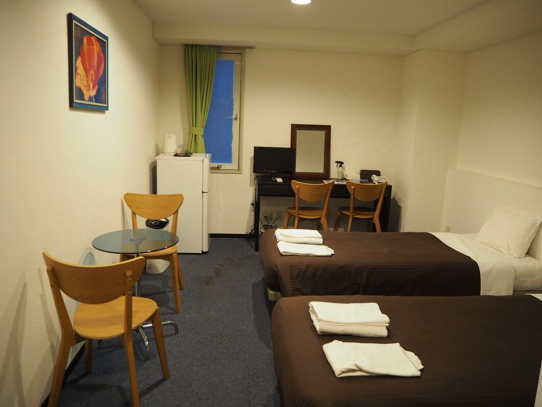 A hotel room with two beds, a desk with a computer, a chair, a small table, and a window with curtains.