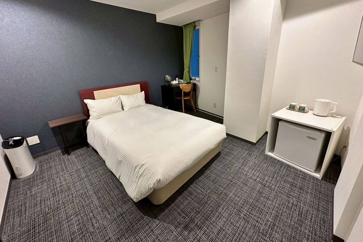 Une chambre d’hôtel comprenant un lit, une table de chevet, un petit bureau avec une chaise, une poubelle et un mini-réfrigérateur.