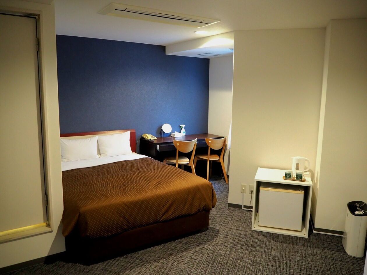 A hotel room with a bed, a desk with a chair, a small table with a coffee maker, and a waste bin.