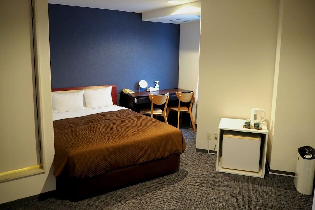 Une chambre d’hôtel comprenant un lit, un bureau avec une chaise, une petite table avec une machine à café et une poubelle.