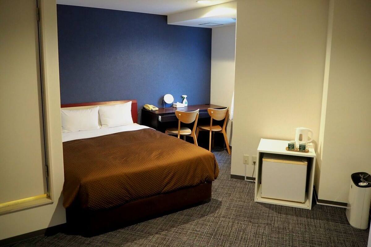 A hotel room with a bed, a desk with a chair, a small table with a coffee maker, and a waste bin.