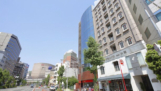 Une rue moderne en milieu urbain, bordée d’immeubles de grande hauteur, dont un imposant gratte-ciel en verre et un bâtiment traditionnel à plusieurs étages, orné d’enseignes.