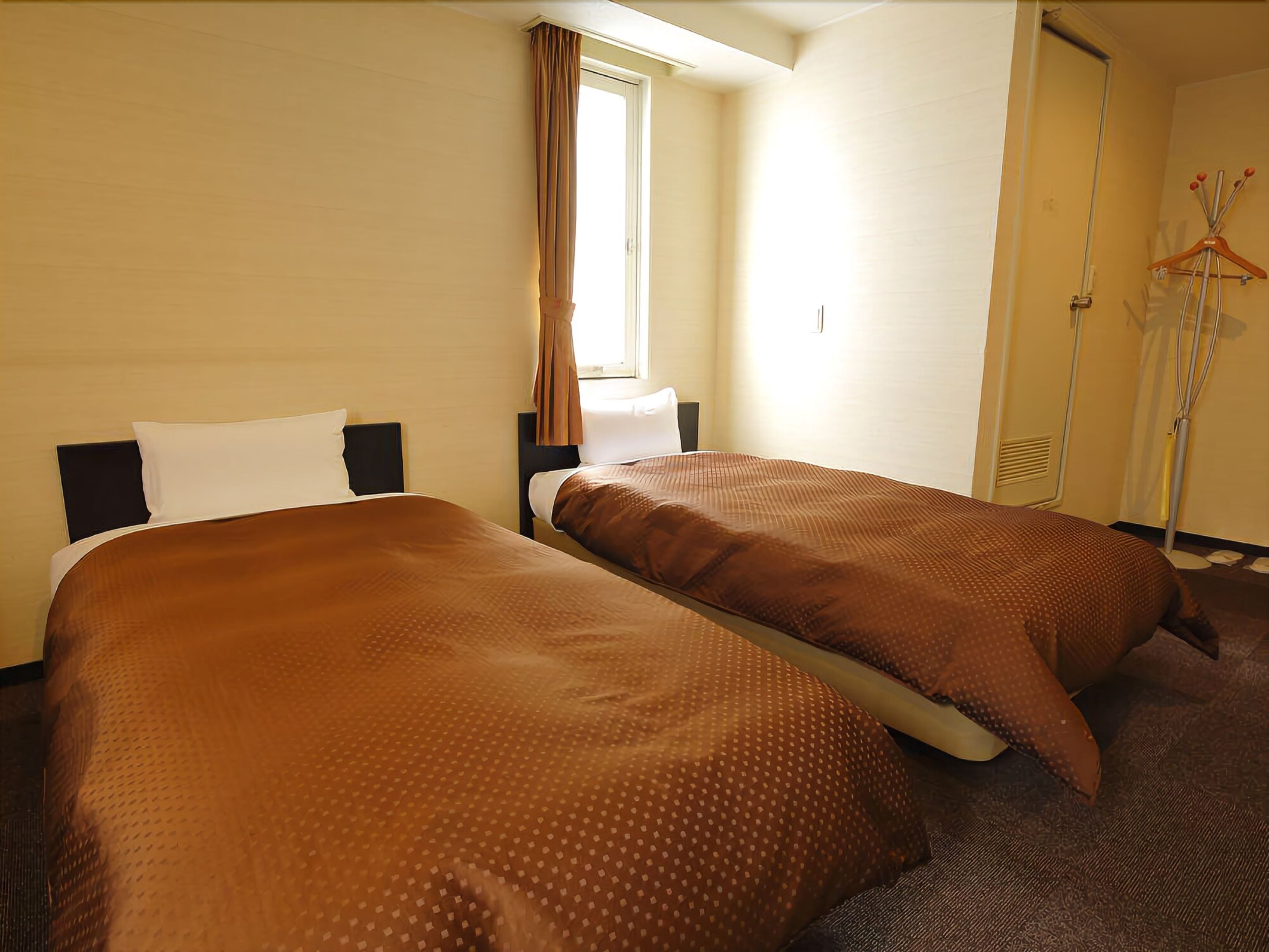 A hotel room with two beds, a window with curtains, a coat rack, and a door.