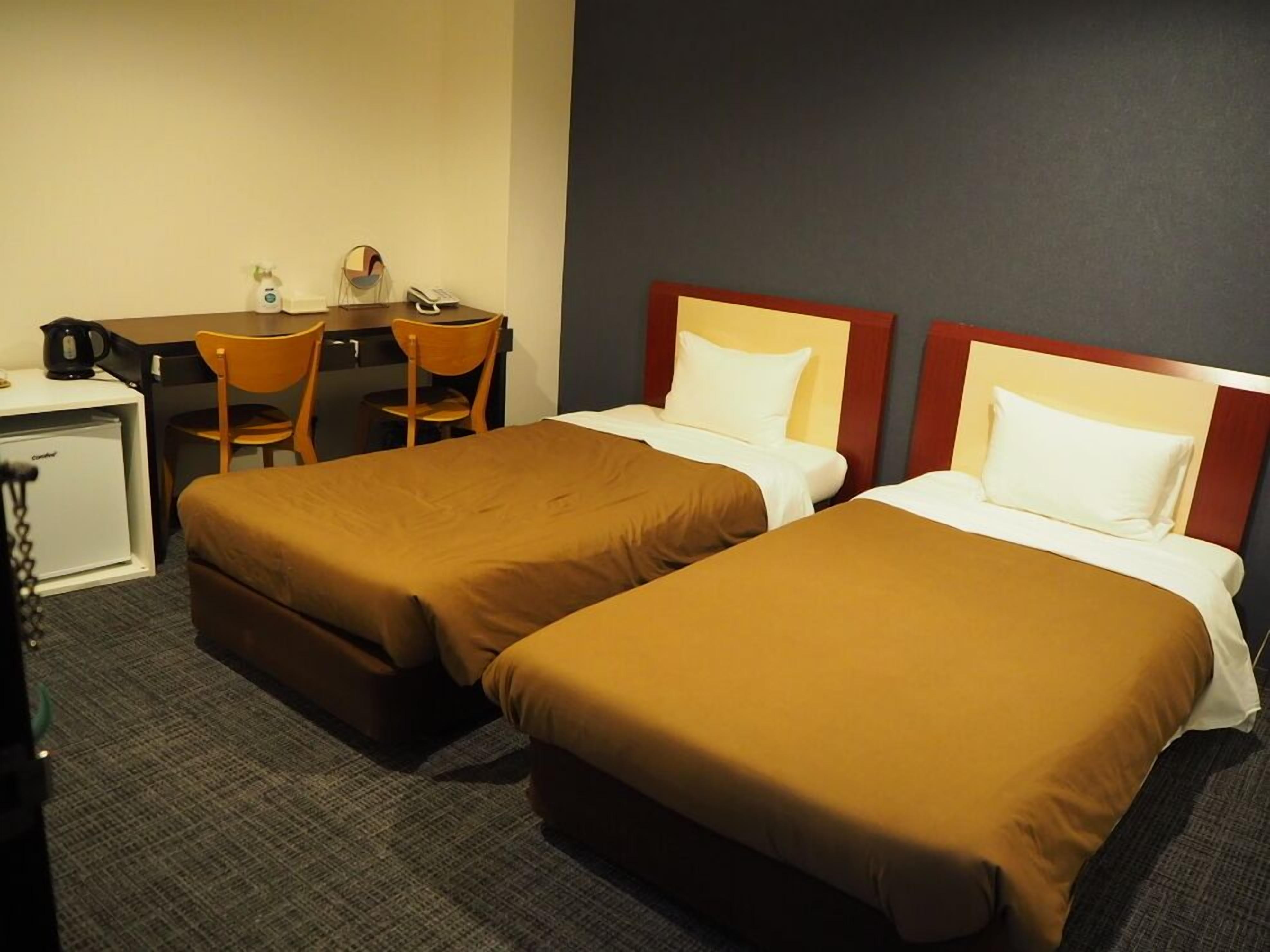 A hotel room with two beds, a desk, a chair, a small refrigerator, and a wall-mounted telephone.