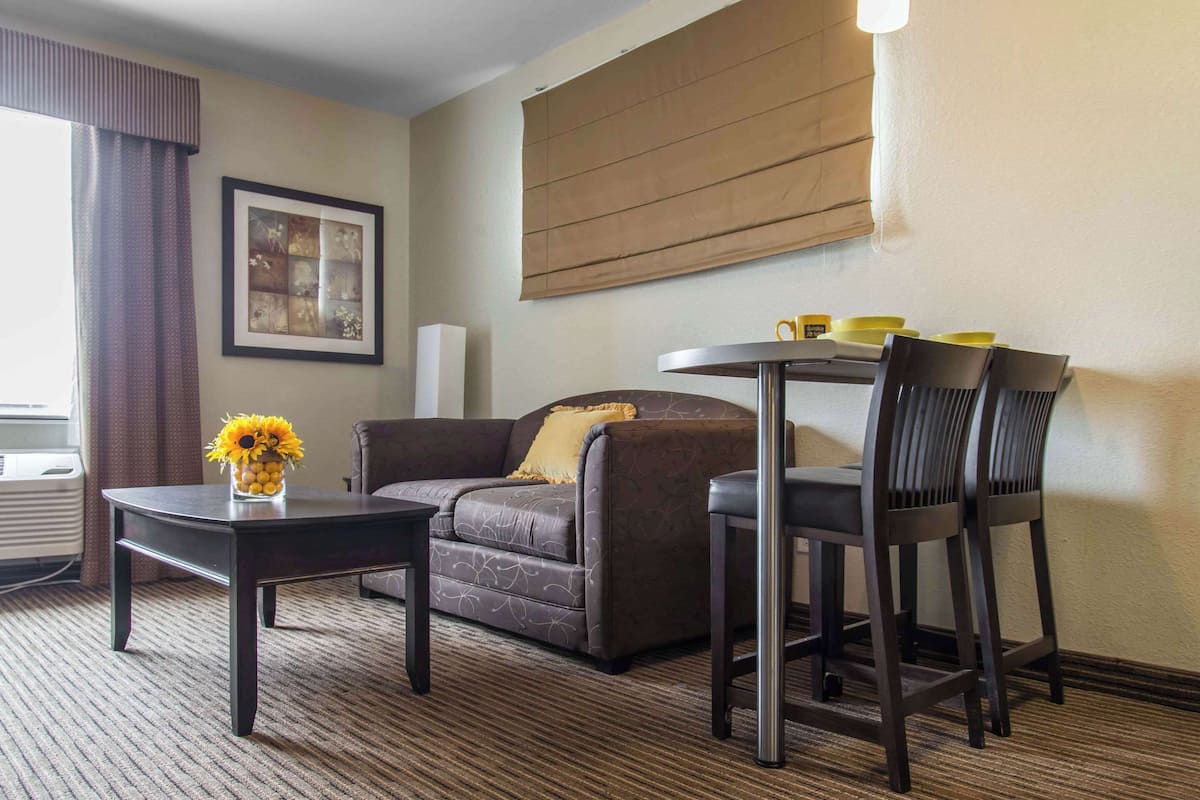 Ein Hotelzimmer mit einer Couch, einem kleinen Tisch mit Sonnenblumen in einer Vase, einem runden Tisch mit Stühlen und einem großen Fenster mit Vorhängen.