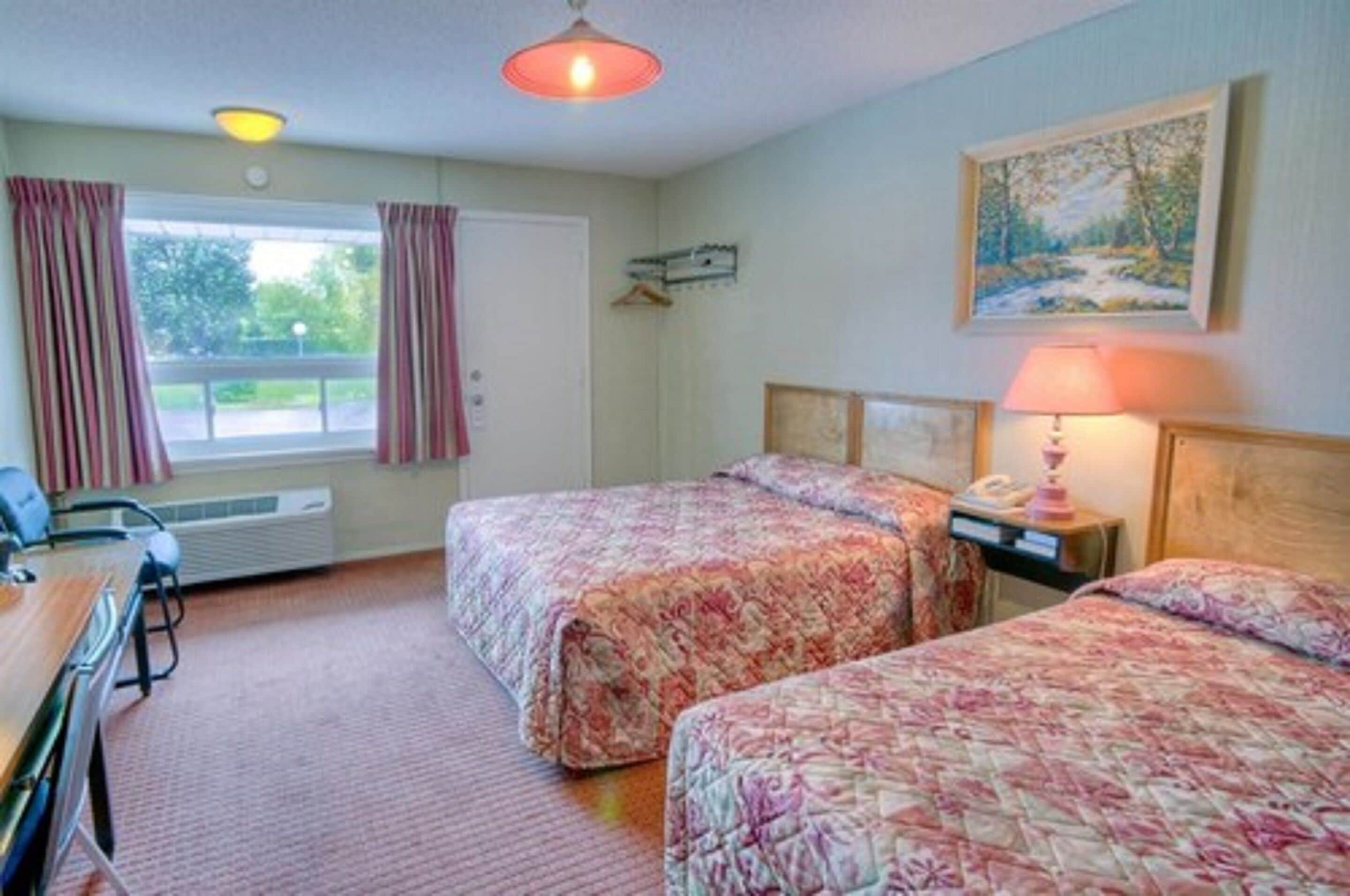 A hotel room with two beds, a desk, a chair, a lamp, and a painting on the wall.
