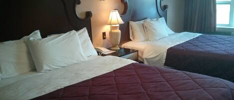 A hotel room with two beds, each with white linens and a dark red quilt. There is a bedside lamp on the left bed, and a phone on the nightstand.