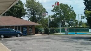 Ein Parkplatz mit einem Gebäude, das eine Schilder für 'Cold Drinks' und einen Geldautomaten zeigt. Vor der Fassade befindet sich eine Fahnenstange mit der US-amerikanischen und der kanadischen Flagge.