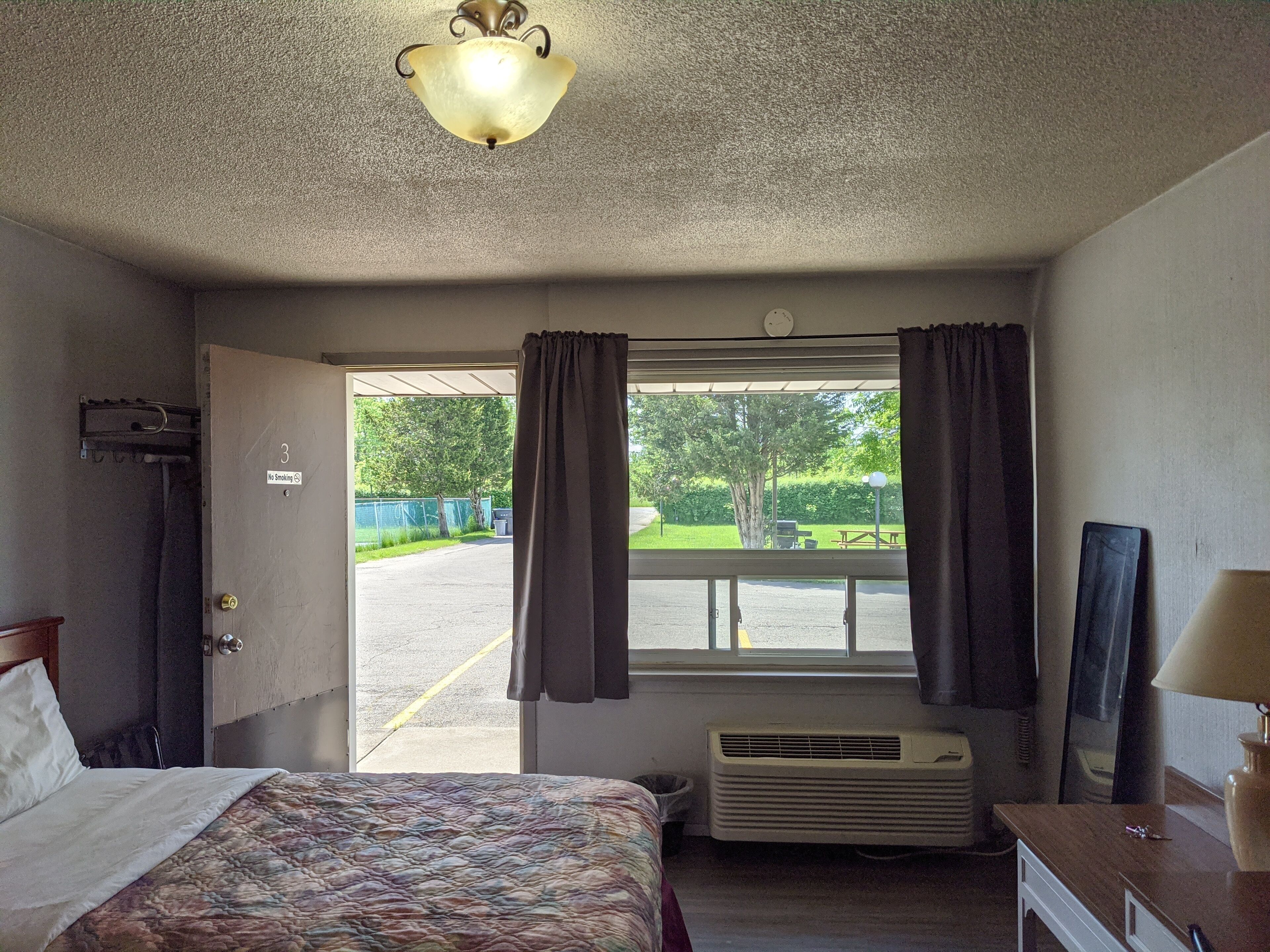 Habitación de motel con cama, ventana con cortinas, lámpara de techo, calentador en la pared y vista al estacionamiento y la vegetación exterior.
