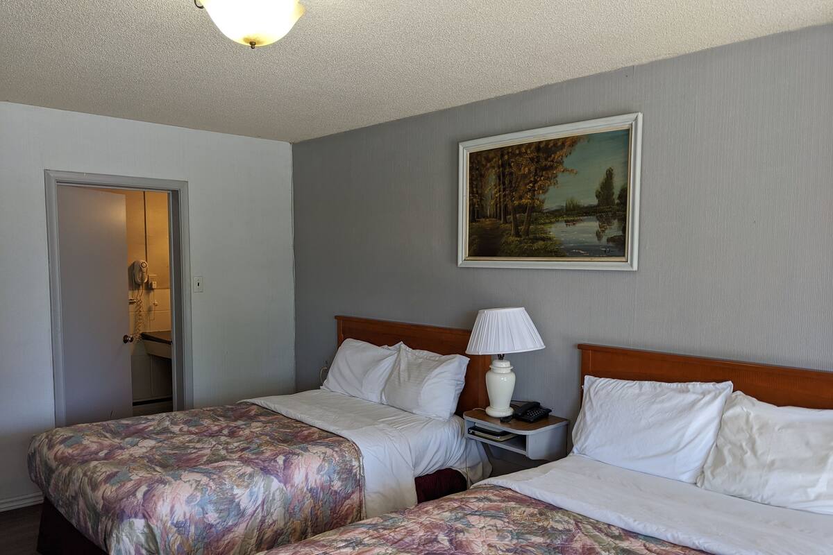 A hotel room with two beds, a painting on the wall, a bedside table with a lamp, and a door leading to a bathroom.