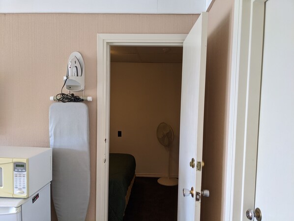 A hotel room with a bed, a bedside table with a microwave, a fan, and a door leading to another room.
