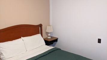 Una cama bien hecha con cabecera de madera, colcha verde y almohada blanca. Se observa una mesita de noche con una lámpara y un enchufe en la pared.