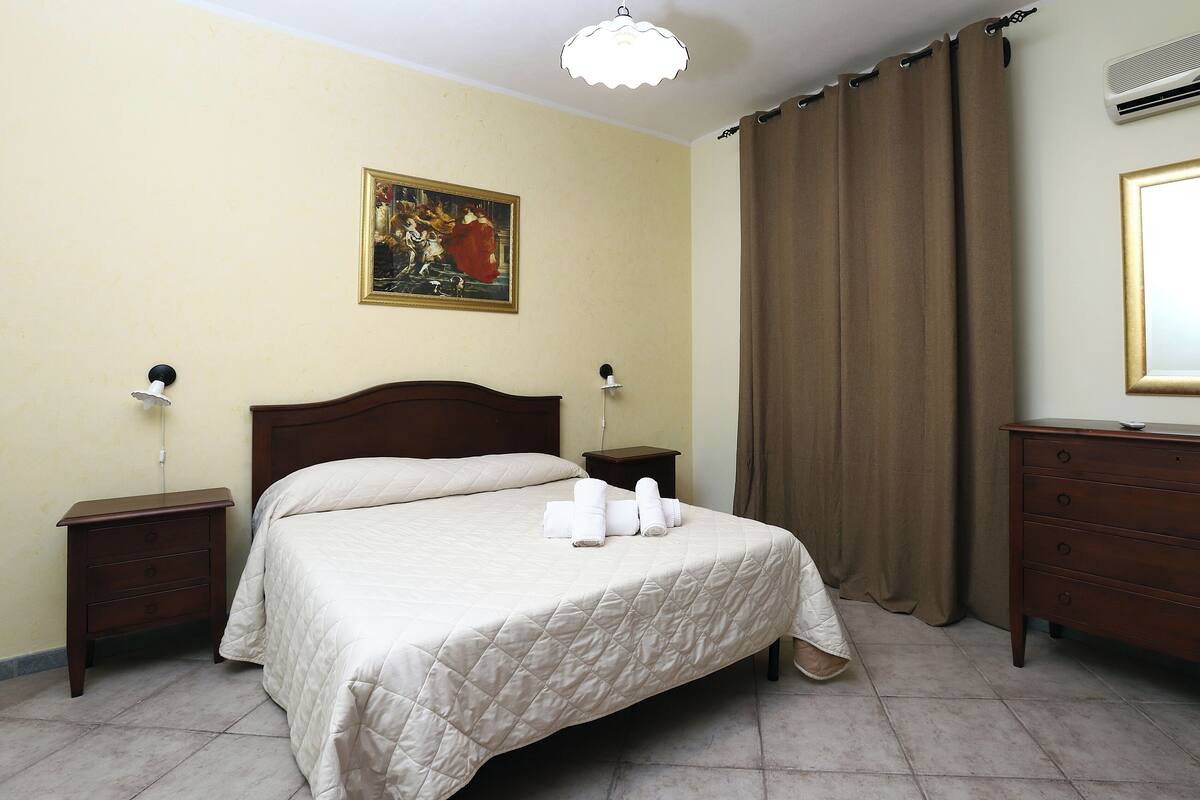 A hotel room with a bed, a nightstand, a dresser, a painting on the wall, and a window with curtains.