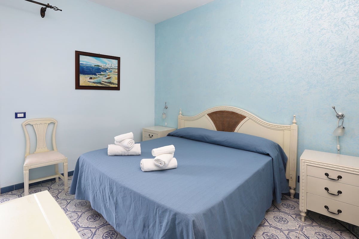 A bedroom with a bed, a chair, a nightstand, a painting, and a window.