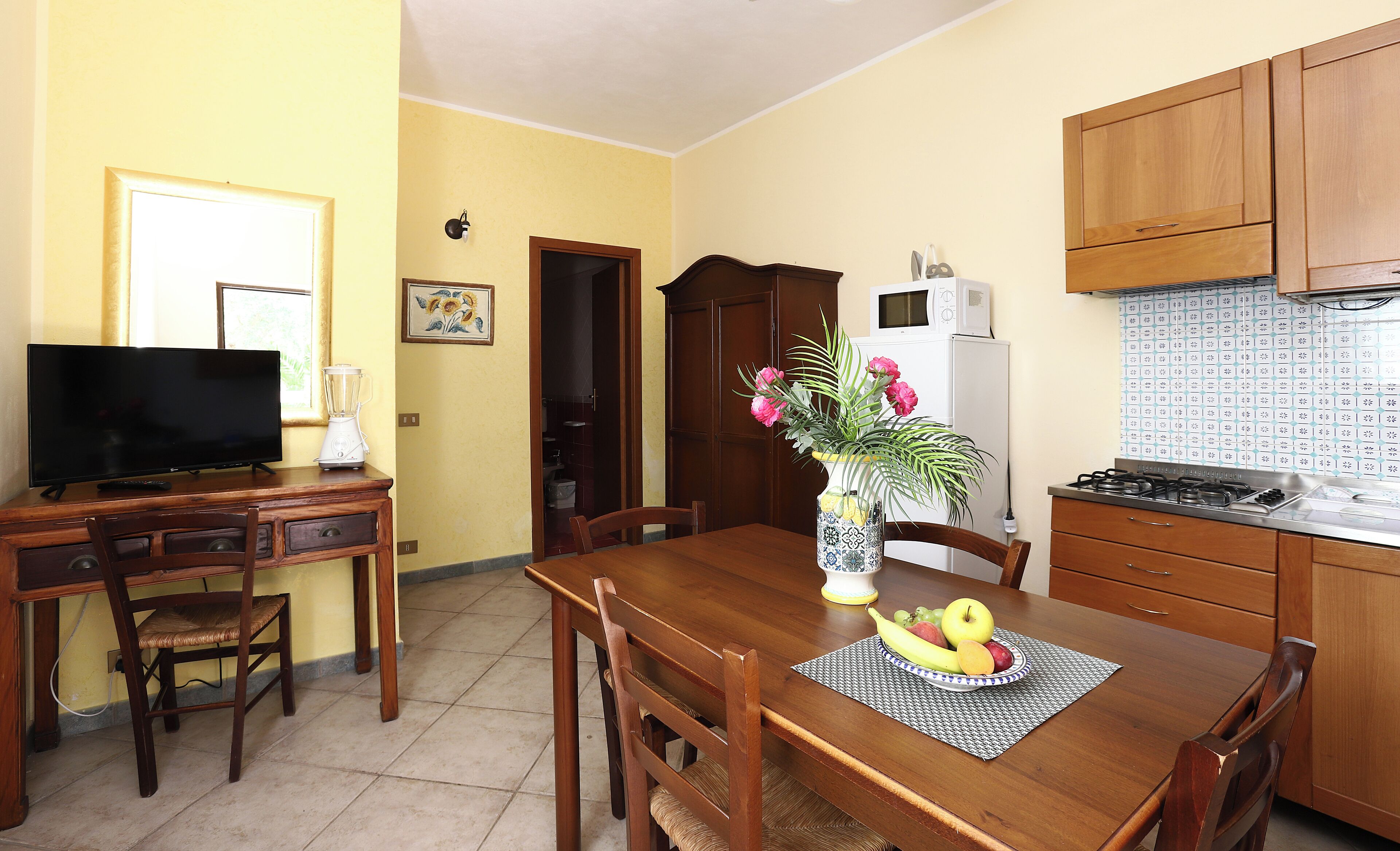 A dining area with a wooden table, chairs, a TV, a blender, and a kitchen with cabinets and a microwave.